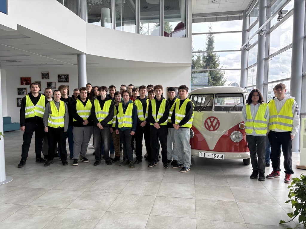 Grupa uczniów ZSEE w żółtych kamizelkach odblaskowych pozująca do zdjęcia podczas wycieczki edukacyjnej. Obok uczniów stoi zabytkowy Volkswagen Transporter T1 z 1964 roku (biało-czerwony) wystawiony w holu nowoczesnego zakładu przemysłowego.